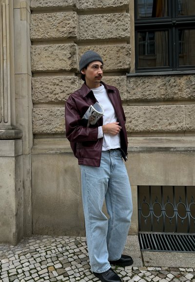 Chaqueta de cuero burdeos, camiseta blanca, jeans de mezclilla azul claro, botas negras. Sosteniendo una revista, de pie contra una pared de piedra texturizada.