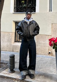 Veste bomber en cuir noir sur un hoodie gris, jeans bleu surdimensionnés avec des poignets retroussés, et chaussures noires. L'arrière-plan présente une fenêtre décorative et une plante.