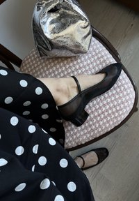Balerini negre din plasă, cu toc pătrat lucios. O curea subțire peste picior. Rochie cu buline negre și albe și o geantă metalizată argintie în fundal.