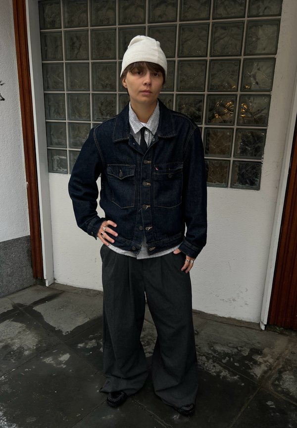 Persoană cu căciulă albă, jachetă din denim, cămașă în carouri și pantaloni largi închiși la culoare stă în interior pe podea cu gresie, sprijinită de un perete din blocuri de sticlă.