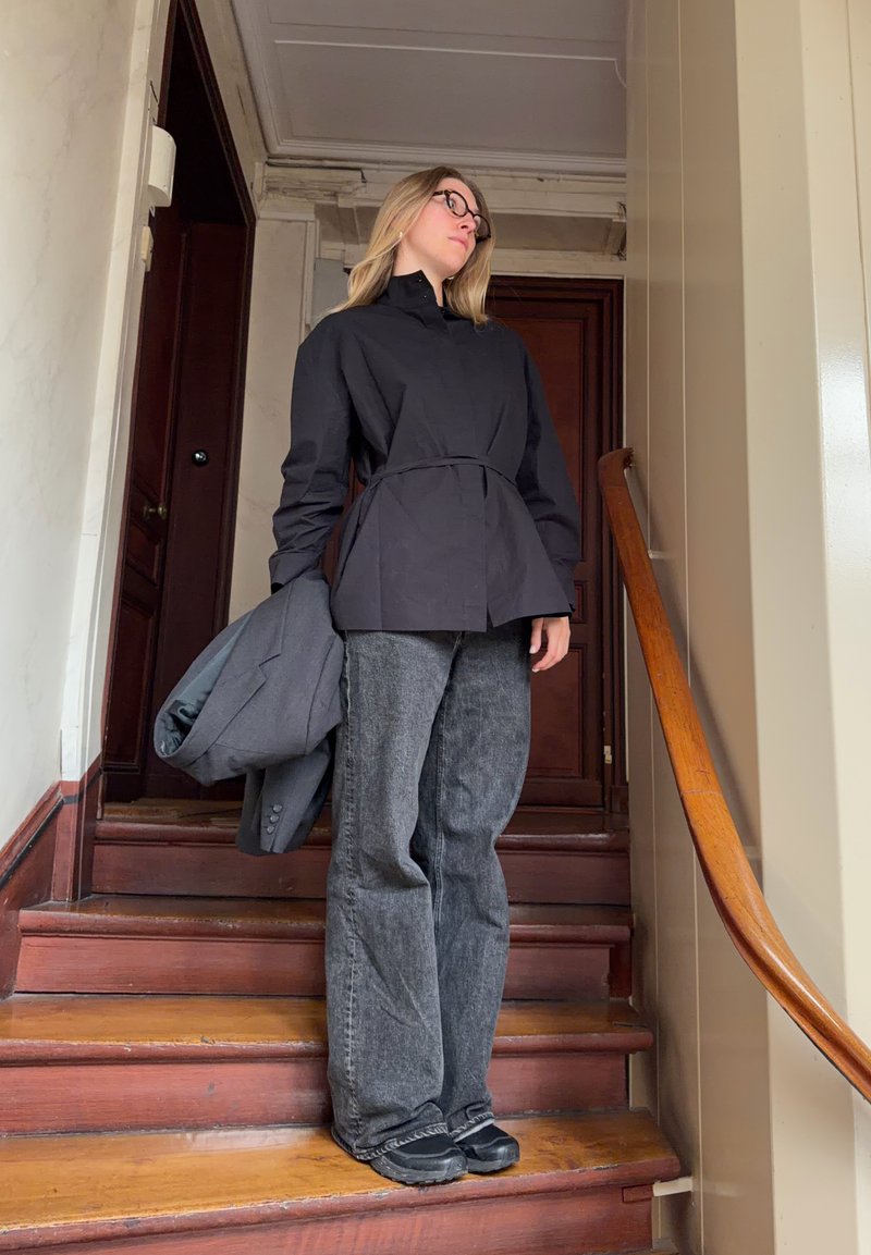 Jeune femme portant une veste noire et un jean gris, debout sur un escalier en bois, tenant un blazer gris, avec une rampe à sa droite.