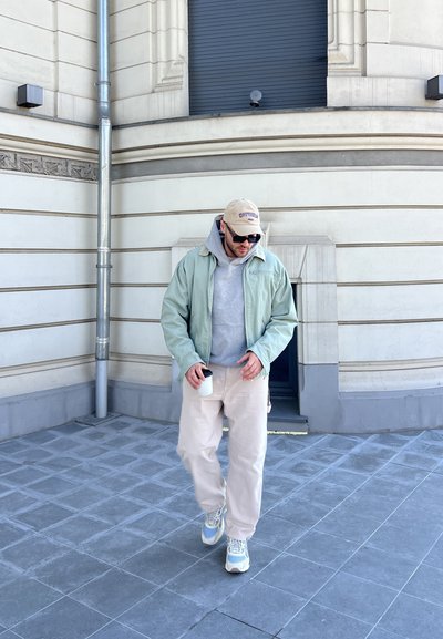 Chaqueta verde claro sobre sudadera gris, pantalones color crema y zapatillas gris-azul. Sosteniendo una taza blanca mientras camina sobre una superficie de azulejos al aire libre.