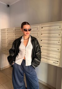 Γυναίκα με γυαλιά ηλίου, μαύρο δερμάτινο μπουφάν, λευκό πουκάμισο με κουμπιά και μπλε παντελόνι, στέκεται με τα χέρια στις τσέπες κοντά σε μπεζ ταχυδρομικούς κουτιούς.