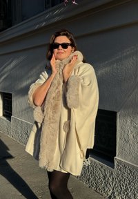 Crèmekleurige cape met nepbontafwerking, ronde vorm en pompondetails. De stoftextuur lijkt zacht en wordt gedragen over zwarte kleding.