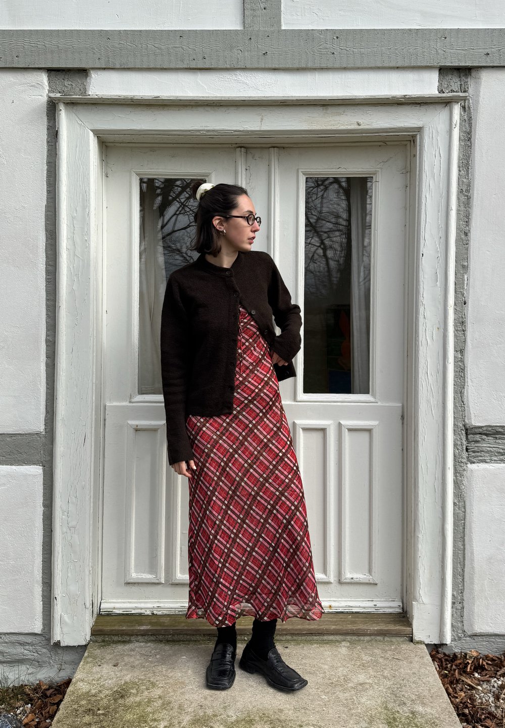Cardigã de malha castanho, vestido maxi xadrez vermelho, meias pretas e sapatos. De pé ao lado de uma porta dupla branca com moldura cinza. Ambiente natural visível.