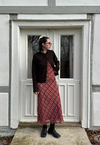 Cardigã de malha castanho, vestido maxi xadrez vermelho, meias pretas e sapatos. De pé ao lado de uma porta dupla branca com moldura cinza. Ambiente natural visível.