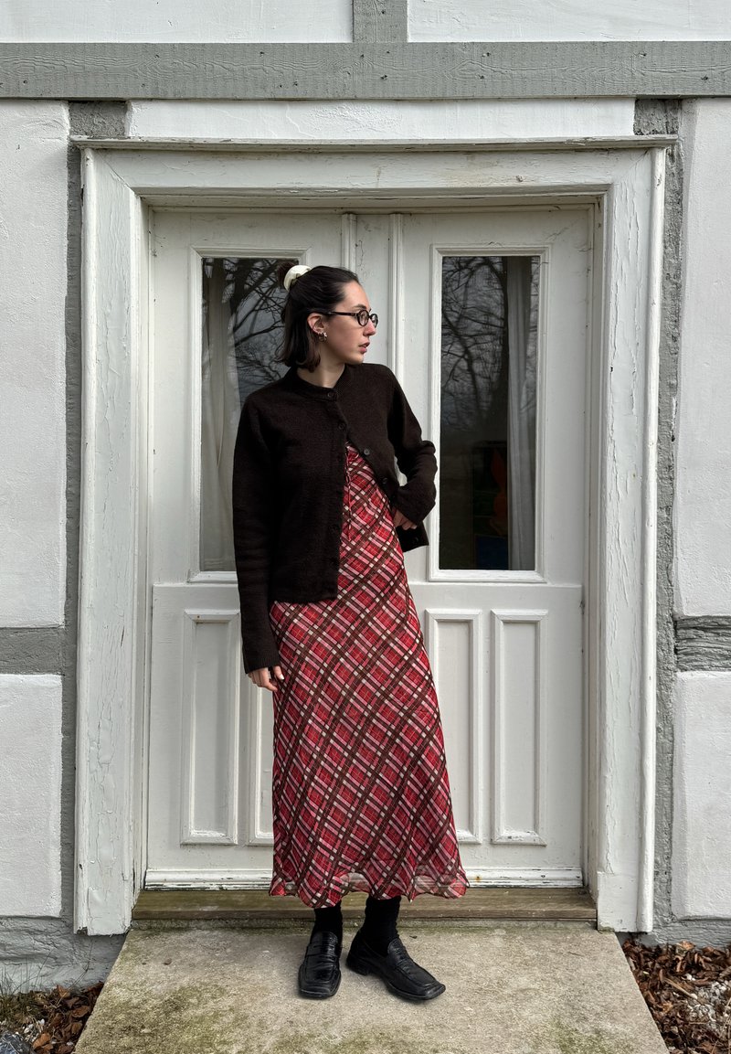 Cardigã de malha castanho, vestido maxi xadrez vermelho, meias pretas e sapatos. De pé ao lado de uma porta dupla branca com moldura cinza. Ambiente natural visível.
