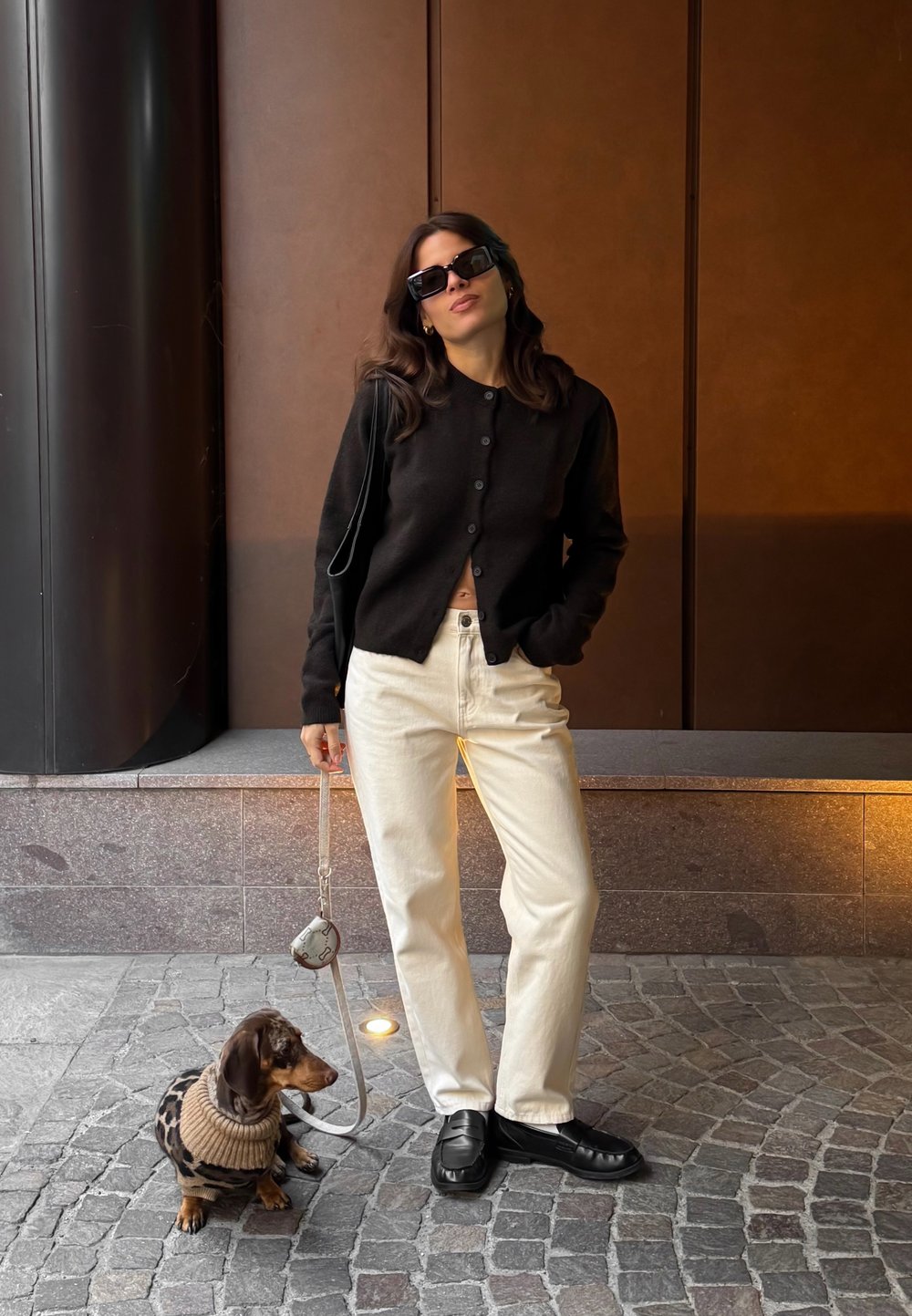 Sort cardigan med knapper foran, hvide jeans og sorte sko. Brun dachshund i en strikket leopardmønstret sweater sidder på brosten.