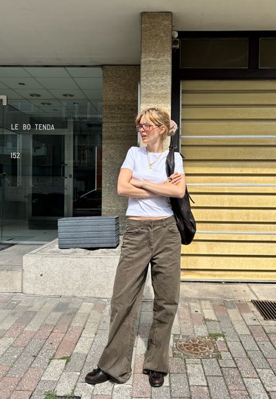Camisa blanca de manga corta, pantalones de pierna ancha verde oliva, zapatos oscuros y bolso negro; de pie frente a un edificio con un fondo a rayas amarillas.