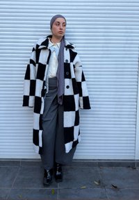 Abrigo de forro polar blanco y negro a cuadros con botones grandes, bufanda gris, camisa blanca texturizada y pantalones grises, combinados con botines negros brillantes.