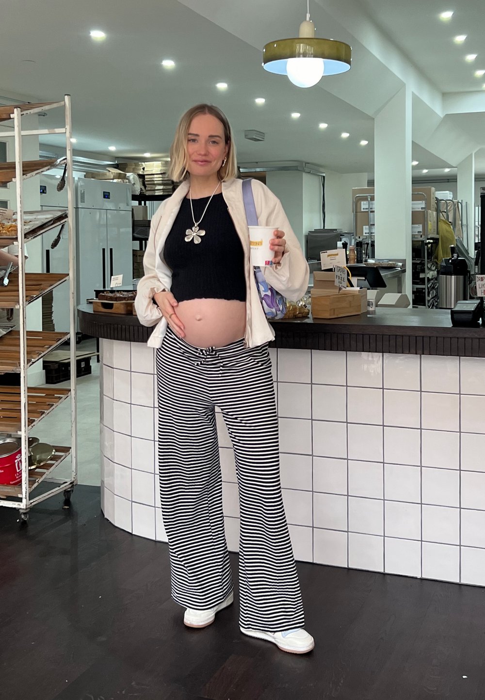 Ένα έγκυο άτομο φοράει μια μαύρη ριμπ μπλούζα, μια λευκή φαρδιά ζακέτα και μαύρο-άσπρες ριγέ παντελόνες, κρατώντας μια κούπα σε ένα καφέ.