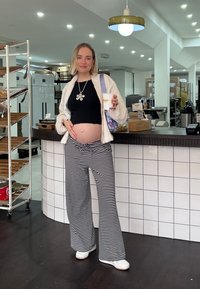 Ένα έγκυο άτομο φοράει μια μαύρη ριμπ μπλούζα, μια λευκή φαρδιά ζακέτα και μαύρο-άσπρες ριγέ παντελόνες, κρατώντας μια κούπα σε ένα καφέ.