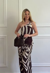 Mujer con el pelo largo y rubio que lleva un vestido de un solo hombro con estampado de cebra, sosteniendo un pequeño bolso negro tejido contra un fondo de pared blanca.