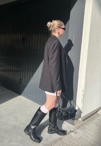 Μαύρο σακάκι, λευκές βερμούδες, δερμάτινες μπότες μέχρι το γόνατο σε μαύρο χρώμα με αγκράφ�ες, λευκές κάλτσες και μια μικρή μαύρη τσάντα. Σκυρόδετο φόντο.