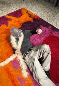 A person lies on a colourful, shaggy rug with orange, purple, and red hues, wearing a grey hat and black clothing, draped in a white faux fur.
