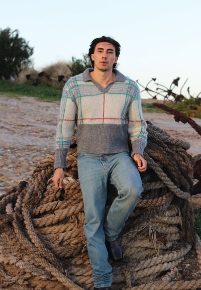 Suéter texturizado en gris, azul y crema con escote en V y patrón de cuadros. Llevado con jeans azul claro y sosteniendo gafas de sol. Sentado sobre cuerda.