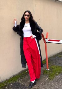 Femme portant un manteau en cuir noir bordé de fourrure, un haut blanc ajusté, un pantalon de survêtement rouge, des mocassins noirs et des lunettes de soleil, appuyée contre un mur beige à l'extérieur.