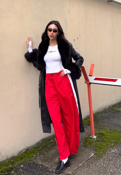 Mujer con abrigo de cuero negro con ribete de piel, camiseta blanca ajustada, pantalones deportivos rojos, mocasines negros y gafas de sol, apoyada en una pared beige al aire libre.