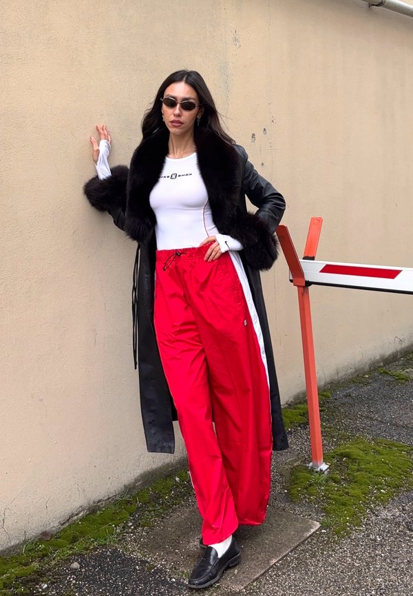 Femme portant un manteau en cuir noir bordé de fourrure, un haut blanc ajusté, un pantalon de survêtement rouge, des mocassins noirs et des lunettes de soleil, appuyée contre un mur beige à l'extérieur.