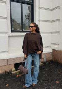 Donna con poncho marrone, jeans blu a gamba larga, scarpe marroni e occhiali da sole, che tiene una borsa marrone, in piedi accanto a un edificio bianco con una finestra.