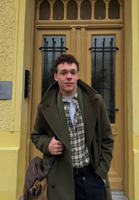 Ciemnozielony wełniany płaszcz na beżowo-czarnej koszuli w kratę, w połączeniu z granatowymi spodniami. Trzyma brązową skórzaną torbę na ramię. Sceneria na zewnątrz.