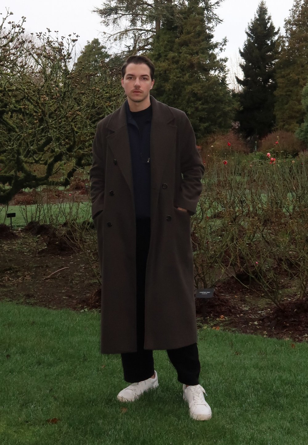 Manteau marron croisé porté sur un pull sombre, pantalon noir et baskets blanches. Debout dans un jardin avec de l'herbe verte et des arbustes.