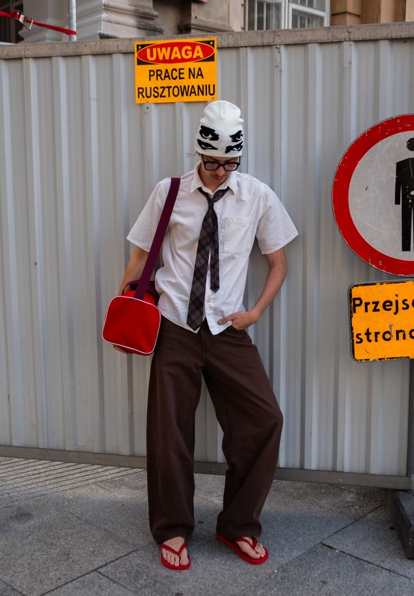 Joven con camisa blanca, corbata oscura, pantalones marrones, chanclas rojas, sombrero blanco con estampado de ojos, sosteniendo una bolsa roja, de pie cerca de una valla metálica de construcción.