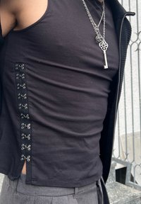 Personne portant un débardeur noir avec des fermetures à crochet argentées sur le côté et des colliers argentés superposés, dont un pendentif en forme de clé.