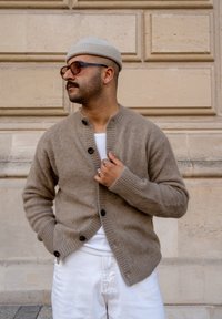 Hnedý vlnený cardigan s rebrovým golierom, čiernymi gombíkmi a dlhými rukávmi, nosený cez biele tričko, skombinovaný s bielymi nohavicami.