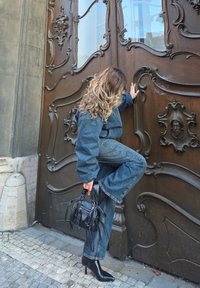 Sinine teksariietus, milles on lahtise lõikega ülaosa ja lehvikpüksid; mustad kontsakingad ja väike käekott; taustal keeruline puidust uks.