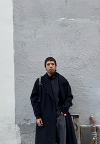 Czarny płaszcz dwurzędowy na czarnej bluzce, szare spodnie o fakturze i torebka z metalowym paskiem; stojąc pod szarym murem.