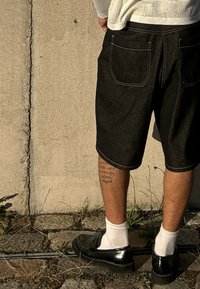 Crne traper šortse s bijelim šavovima, s dva stražnja džepa, nosi se uz crne mokasine i bijele čarape na betonskoj pozadini.