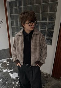 Veste courte à carreaux marron et beige avec fermeture éclair, poignets côtelés, portée sur une chemise noire et un pantalon, debout contre un mur en blocs de verre.
