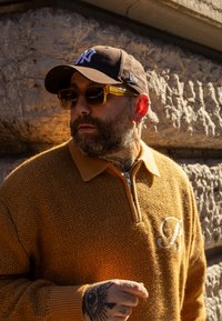 Casquette marron et lunettes à verres jaunes, pull tricoté beige avec fermeture éclair et accent brodé. Tatouage visible sur la main, fond en pierre texturée.