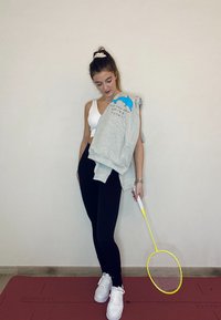 Grey sweatshirt featuring a blue dolphin graphic, black leggings, and white trainers. Holding a yellow badminton racket. Light background.