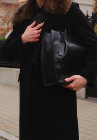 Sort skinnveske med teksturert overflate, glidelåslukking og en tagg. Holdt i én hånd, komplettert med en svart frakk med pelskrage.