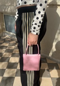 Personne portant un pantalon noir avec des rayures beiges et un haut à manches longues blanc à pois noirs, tenant un petit sac à main rose.