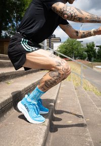 Sapatos de corrida azuis com uma sola branca espessa, combinados com meias de padrão azul claro. A pessoa está a agachar-se em degraus de concreto.