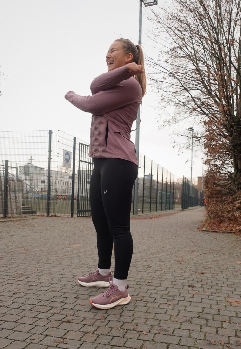 Conjunto atlético deportivo que incluye una chaqueta de cremallera morada clara con paneles texturizados, leggings negros y zapatillas para correr moradas claras en un camino pavimentado.