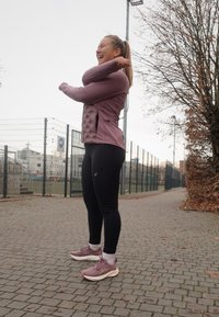 Conjunto atlético deportivo que incluye una chaqueta de cremallera morada clara con paneles texturizados, leggings negros y zapatillas para correr moradas claras en un camino pavimentado.