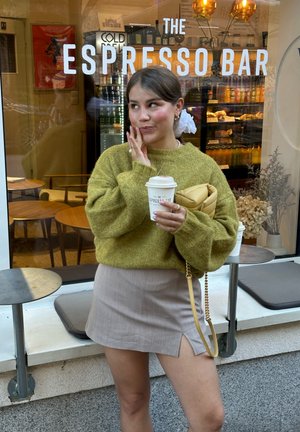 Mujer con suéter verde y falda beige sostiene una taza de café fuera de una barra de espresso, con asientos de café visibles a través de la ventana.