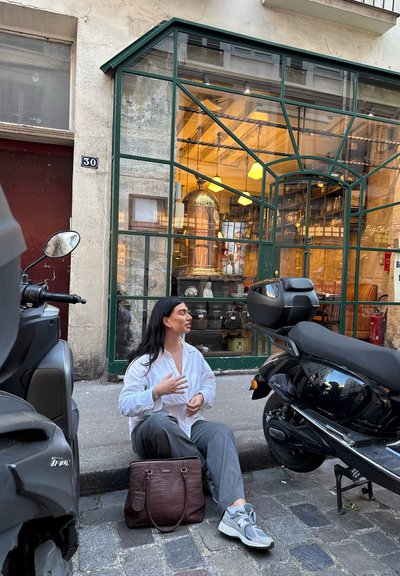 Una moto negra y una moto gris aparcadas al lado de un café con fachada de cristal, interior de madera, calle de adoquines y una bolsa marrón en el suelo.