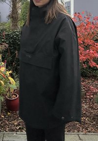 Juodas vandeniui atsparus švarkas su aukšta apykakle, turi priekinę kišenę ir užsegimus, dėvimas virš juodų kelnių sodininkystės aplinkoje.