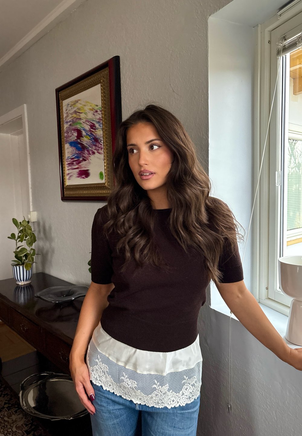 Mujer con cabello largo y ondulado de color castaño, vestida con una blusa oscura con dobladillo de encaje y jeans azules, de pie junto a una ventana en una habitación con una pintura y plantas.