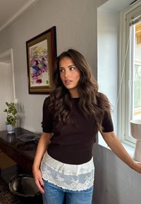 Mujer con cabello largo y ondulado de color castaño, vestida con una blusa oscura con dobladillo de encaje y jeans azules, de pie junto a una ventana en una habitación con una pintura y plantas.