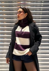 Raidallinen pusero valkoisena ja tummanviolettina, jossa on korkeakaulus ja vetoketjusomistus, yhdistetty mustaan takkiin ja farkkushortseihin.