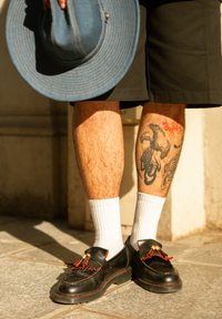 Schwarze Lederschuhe mit roten Akzenten und Quasten, weiße Socken, Tätowierung von Skorpionen am Bein, Denim-Hut in der Hand, trägt dunkle Shorts.
