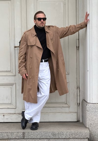 Gabardina beige con grandes solapas, llevada sobre un suéter de cuello alto negro; combinada con pantalones blancos y zapatos negros, sobre un fondo de puerta neutra.