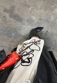 Femme portant des bottes en cuir noir et un manteau, tenant un tissu blanc brodé de fleurs noires et un petit sac rouge avec des clous dorés.