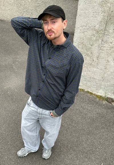 Camisa de cuadros en verde y azul marino, jeans grises, gorra negra y zapatillas grises. Presenta dos bolsillos en el pecho y un corte relajado.